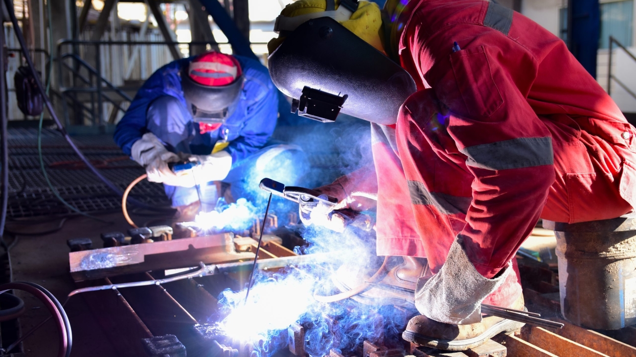 Two welder is assembling the workpiece by process shielded metal arc welding (SMAW). Welder in red uniform, safety shoes, leather gloves, welding mask. He is sitting and welding.