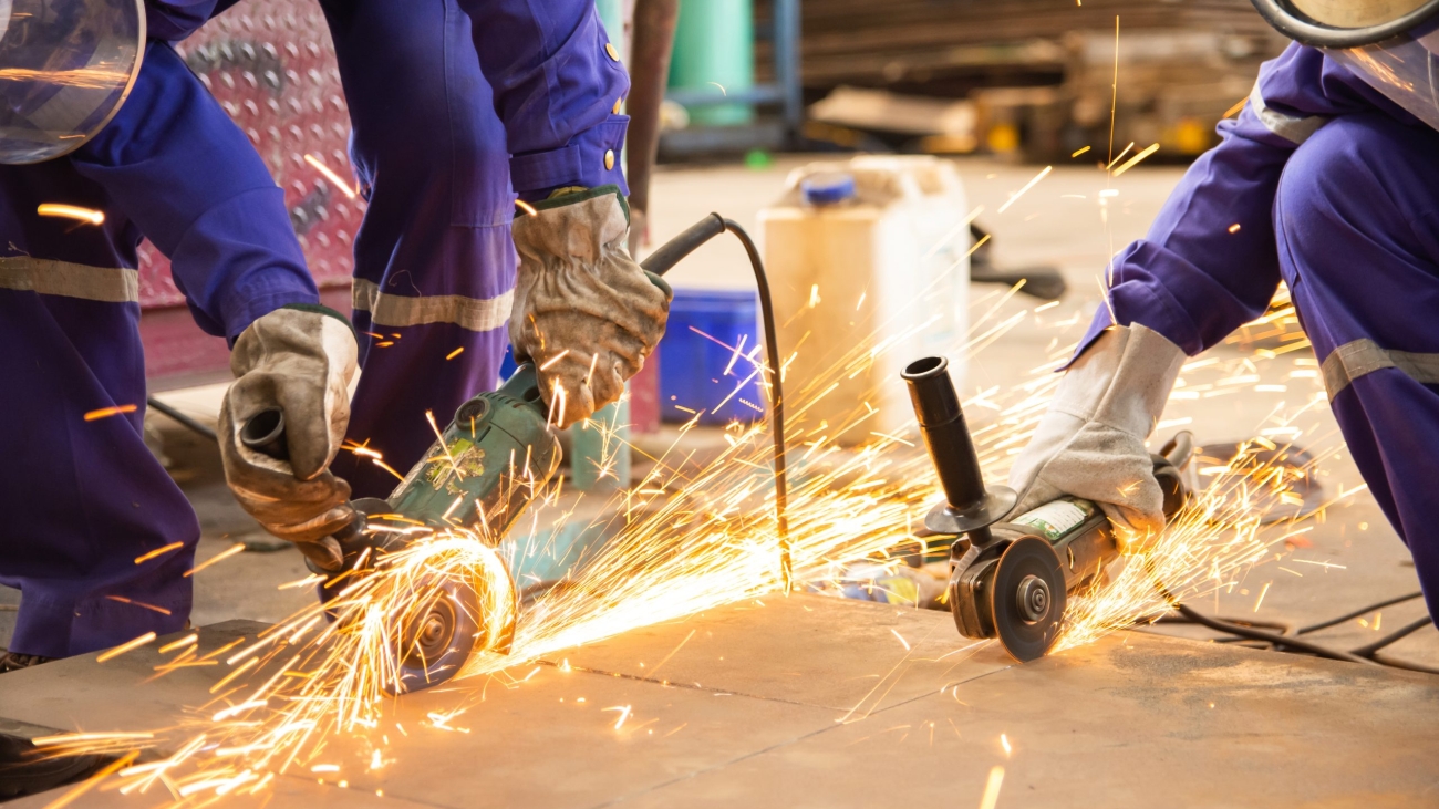 Two male workers cutting metal sheets with electric grinder in the workshop.Mechanic wear personal protective equipment(PPE) while working.Leather gloves and face shield.